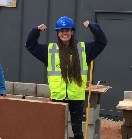 A young woman wears a high vis jacket and a construction helmet. She is doing a strong woman pose. She is building a brick wall.