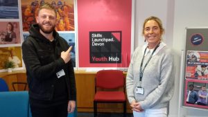 an image on a man (Tom) wearing black trouser and top pointing at the youth hub logo, and a woman (Rhian) dressed in white trousers and jumper.